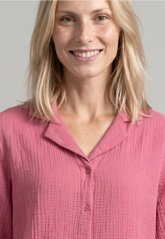 Une femme souriante aux cheveux blonds porte une blouse rose texturée.
