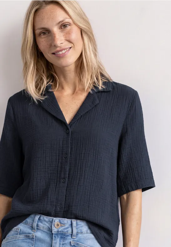 Eine lächelnde Frau mit blonden Haaren trägt eine dunkelblaue Bluse und Jeans.