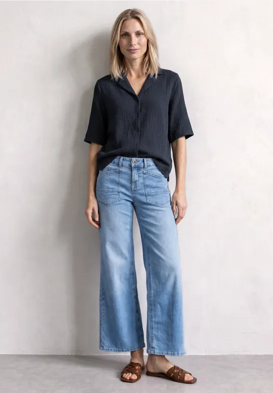 Frau in schwarzer Bluse, weitem Jeans und Sandalen vor neutralem Hintergrund.