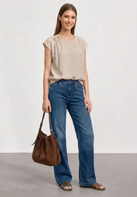 Vrouw met beige top, rechte blauwe jeans, bruine tas en sandalen voor een witte achtergrond.