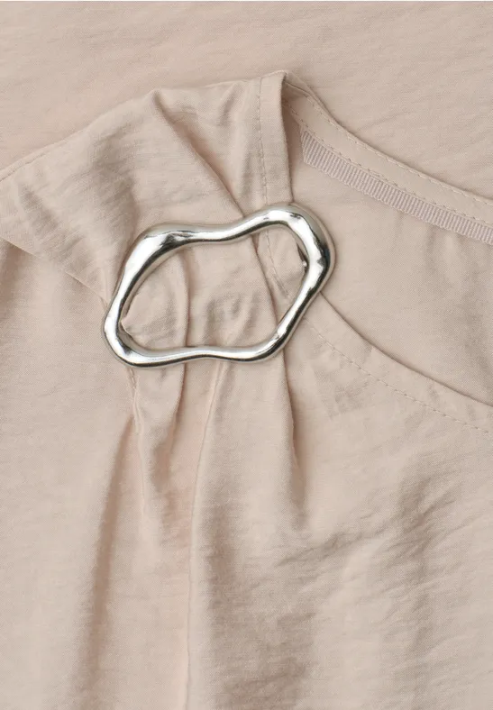 Close-up van een beige top van licht stof met onregelmatige zilverkleurige ring bij de schouder.