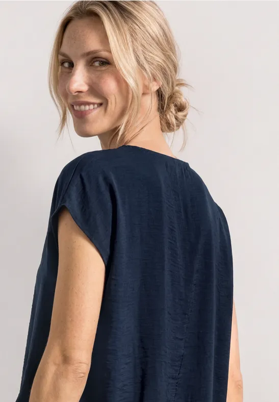 Rugaanzicht van een persoon met blond haar in een lage knot en een donkerblauw, kortmouwig bovenstuk.