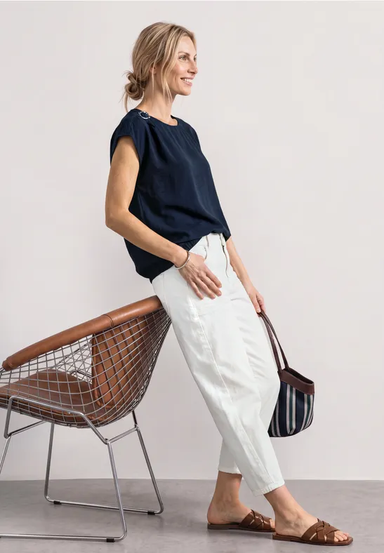 Vrouw leunt nonchalant op stoel, donkerblauw topje, witte broek, bruine sandalen, gestreepte tas.