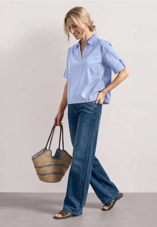 Vrouw in lichtblauw kort overhemd, losse jeans en rieten tas, loopt ontspannen.