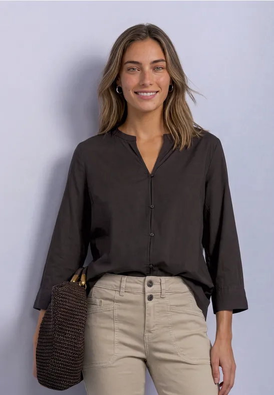 Femme souriante en chemisier noir et pantalon beige, sac en bandoulière, sur fond clair.