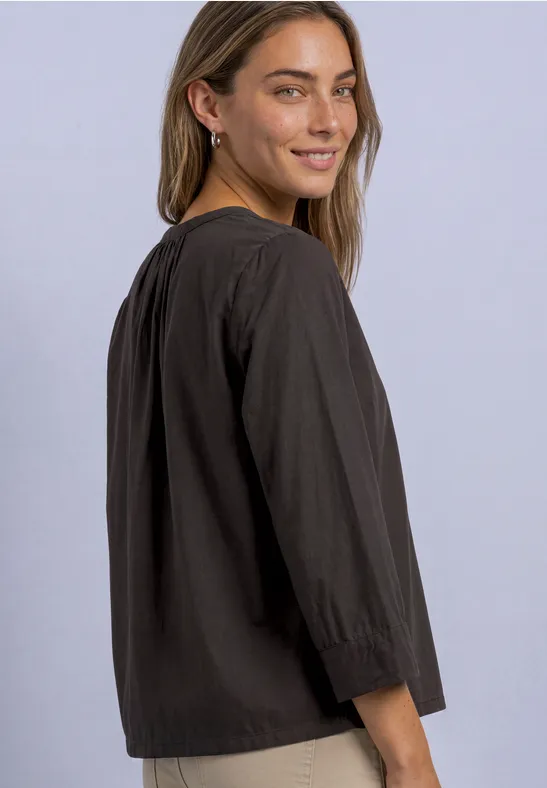Jeune femme en blouse noire ample à manches 3/4, souriant par-dessus l'épaule sur fond clair.