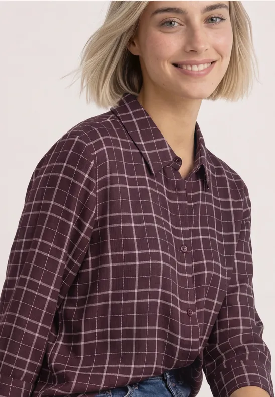 Eine lächelnde Frau mit kurzen blonden Haaren trägt ein dunkelrot-kariertes Hemd.