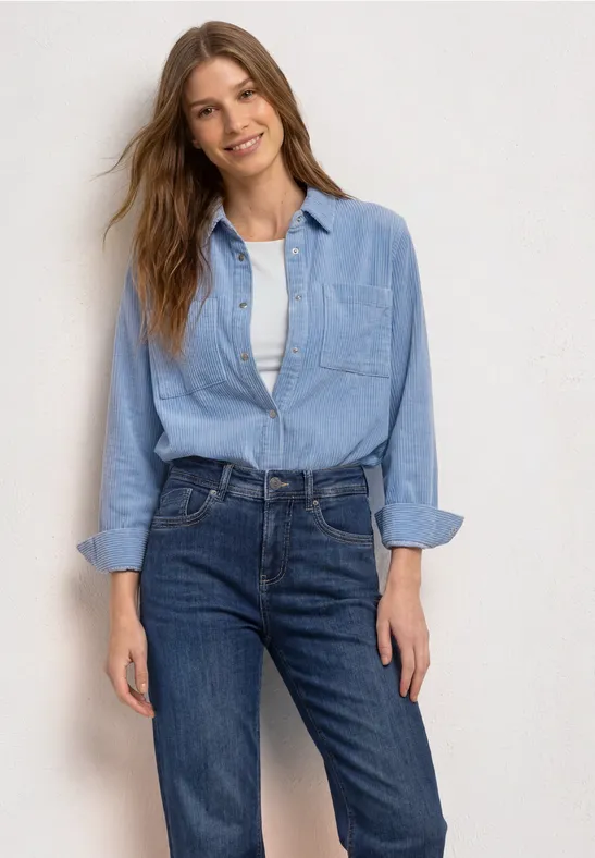 Eine Frau lächelt in Jeanshose und hellblauer Jeansbluse vor hellem Hintergrund.