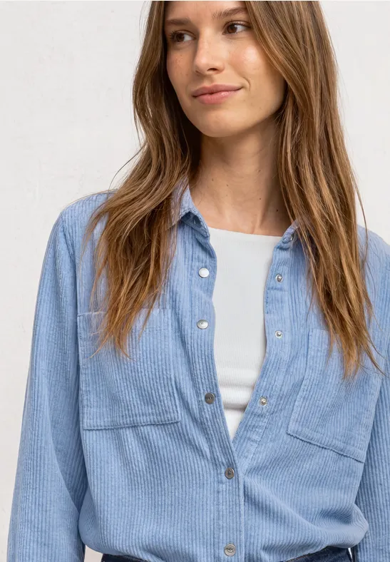 Junge Frau in hellblauem Jeanshemd über weißem Top, leicht lächelnd vor neutralem Hintergrund.