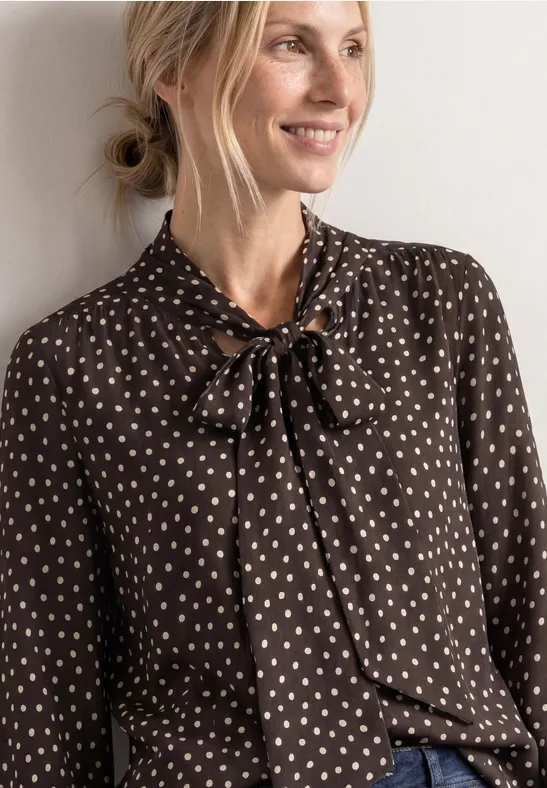 Femme blonde portant un chemisier marron à motifs blancs en forme de cœur et un nœud au col, souriante de profil.