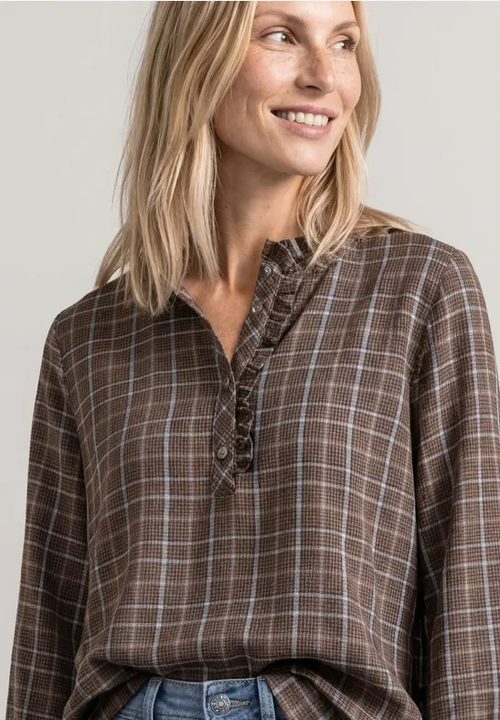 Blonde Frau lächelt seitlich, trägt braun-kariertes Blusenshirt mit Rüschen vor neutralem Hintergrund.