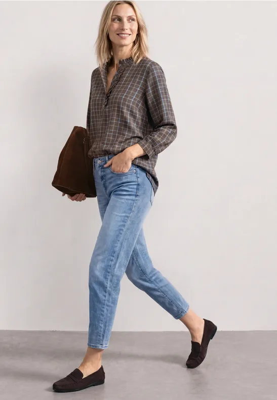 Eine lächelnde Frau in kariertem Hemd, Jeans und Mokassins hält eine braune Tasche.