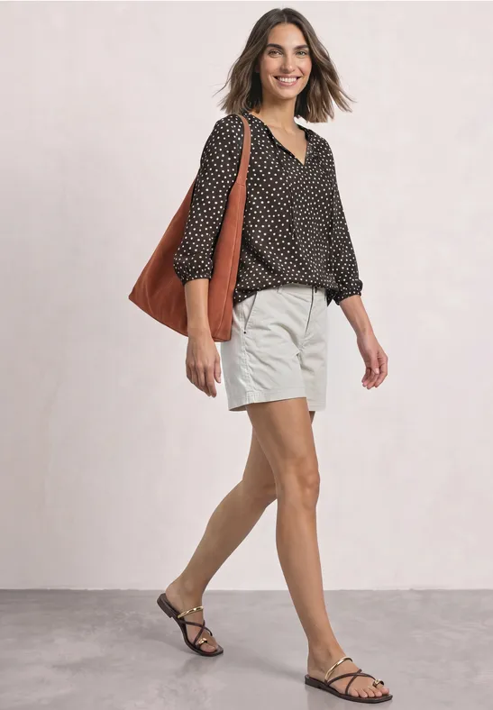 Eine lächelnde Frau in gepunkteter Bluse, beigen Shorts und schwarzen Sandalen trägt eine große Tasche.
