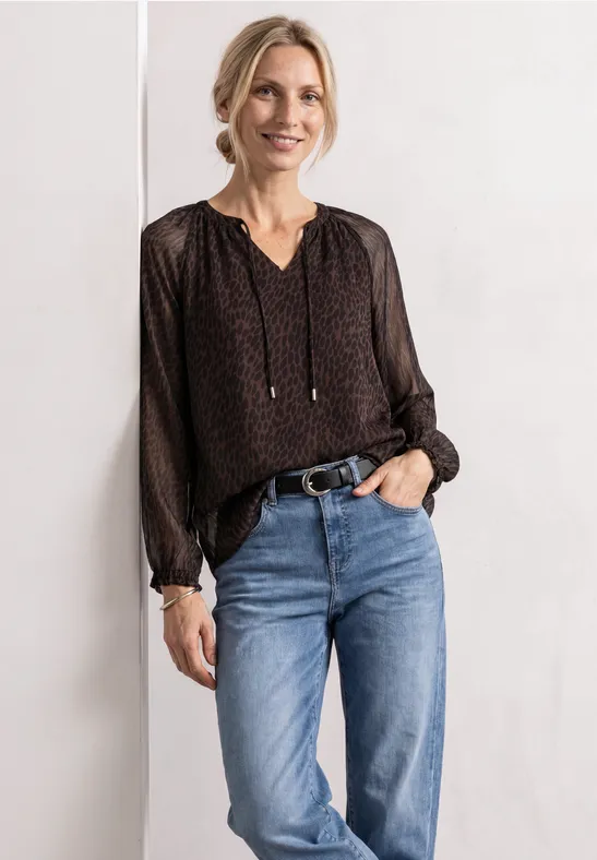 Une femme souriante porte un chemisier noir à pois et un jean bleu devant un fond blanc.