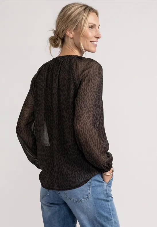 Femme en top noir transparent à motif et jean, souriante, regard par-dessus l’épaule.