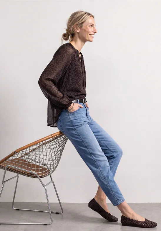 Femme en pull marron et jean bleu, assise de profil sur une chaise filaire en souriant.