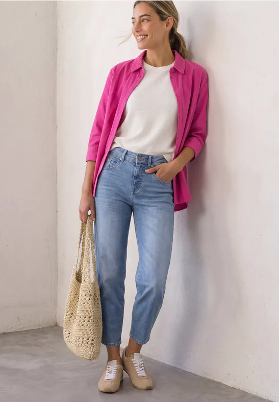 Frau in weißem T-Shirt, pinker Bluse, blauen Jeans und Sneakers lehnt lächelnd an Wand, trägt gehäkelte Tasche.