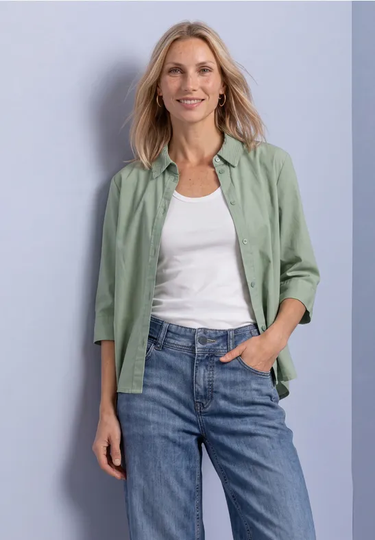 Eine lächelnde Frau mit blondem Haar trägt ein grünes Hemd über einem weißen Shirt und blauen Jeans.