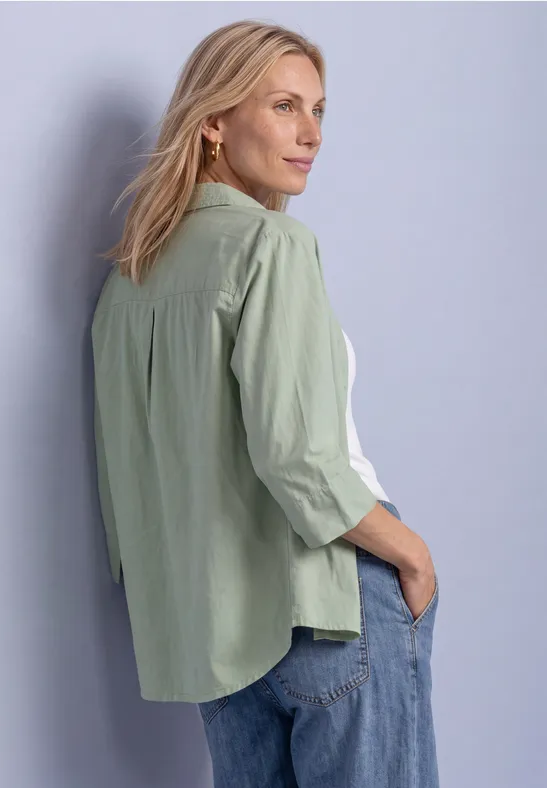 Eine Frau in hellgrüner Bluse und Jeans steht seitlich vor einer Wand und blickt nach rechts.