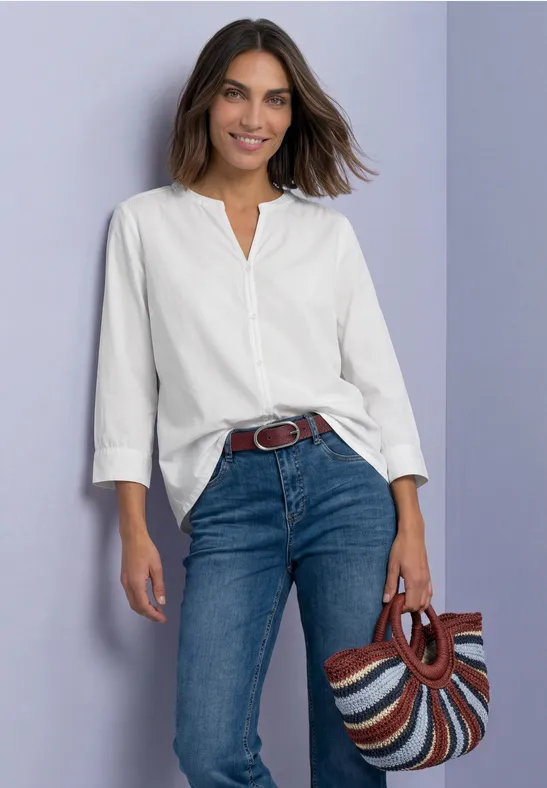 Femme en chemise blanche et jeans bleus, sac rayé multicolore, devant un mur violet, souriante.