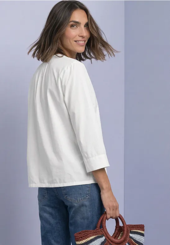 Femme en chemisier blanc et jean, souriante par-dessus l’épaule, tenant un sac aux poignées rondes en bois.