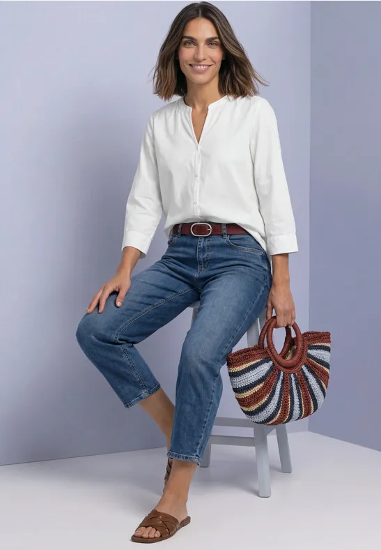 Une femme souriante assise sur un tabouret, blouse blanche, jean, ceinture en cuir et sac paille coloré.