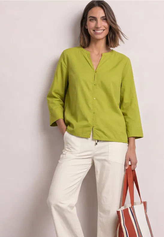 Femme souriante en chemise vert clair et pantalon blanc, tenant un sac rouge et blanc.