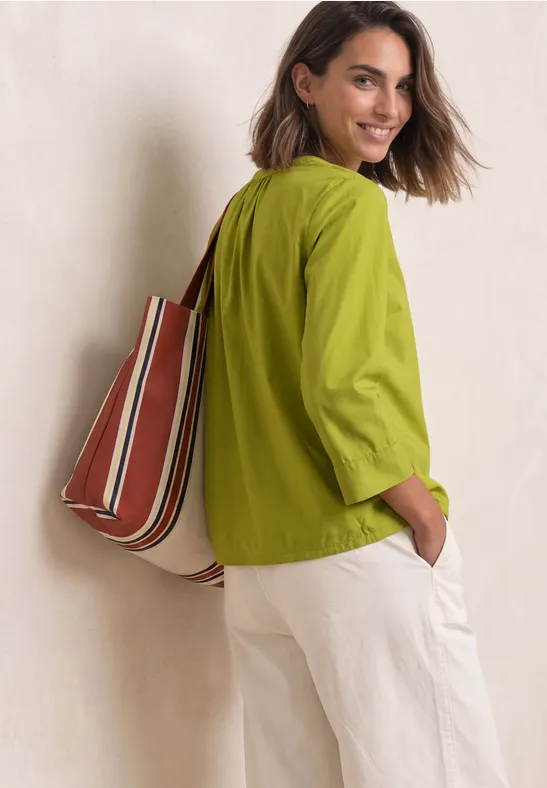 Femme souriante en chemise verte, pantalon blanc, sac rayé porté à l’épaule