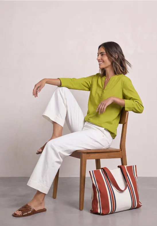 Femme assise sur chaise en bois, chemisier vert, pantalon blanc et sac rayé posé par terre.