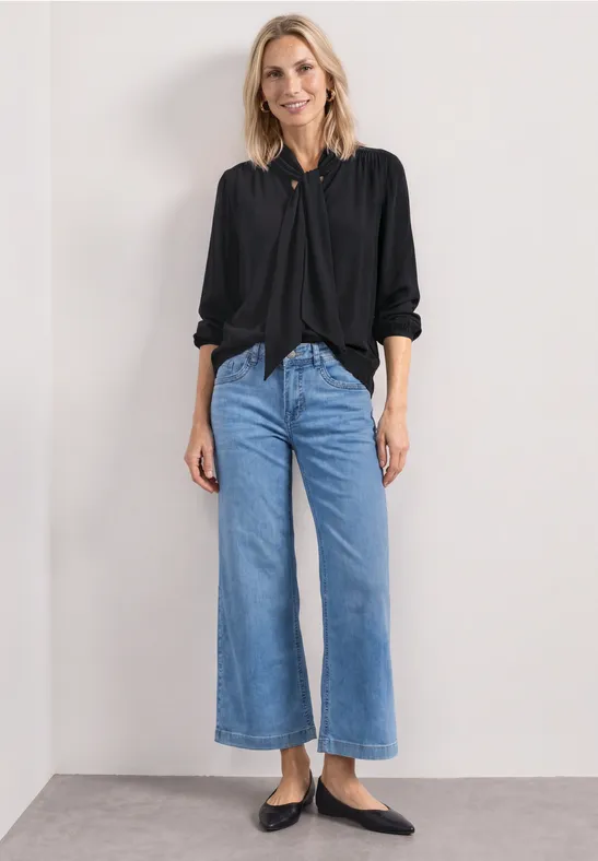 Femme en chemise noire, jean large bleu clair raccourci et ballerines noires devant un mur blanc.