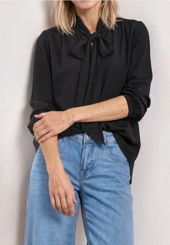 Femme en chemisier noir à col noué et jean bleu, bras croisés devant le buste.