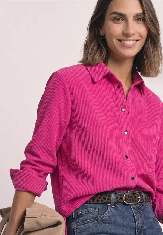 Une femme sourit détendue, vêtue d’une chemise en velours côtelé rose fuchsia, jean et ceinture.