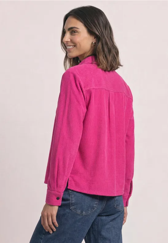 Femme de profil portant une chemise rose et un jean bleu, souriante.