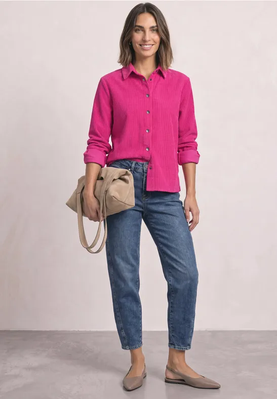 Femme souriante en chemise rose fuchsia, jean bleu, besace beige et ballerines beiges sur fond neutre.