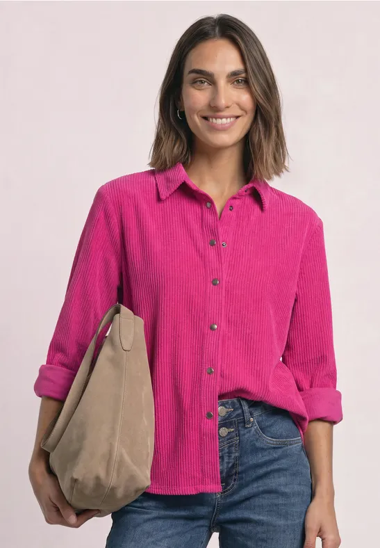 Femme souriante en chemise rose à manches retroussées, jean et sac beige sur l'épaule.
