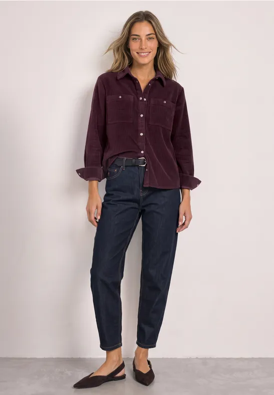 Vrouw in een bordeauxrode corduroy blouse, donkerblauwe jeans en zwarte ballerina's voor een neutrale achtergrond.
