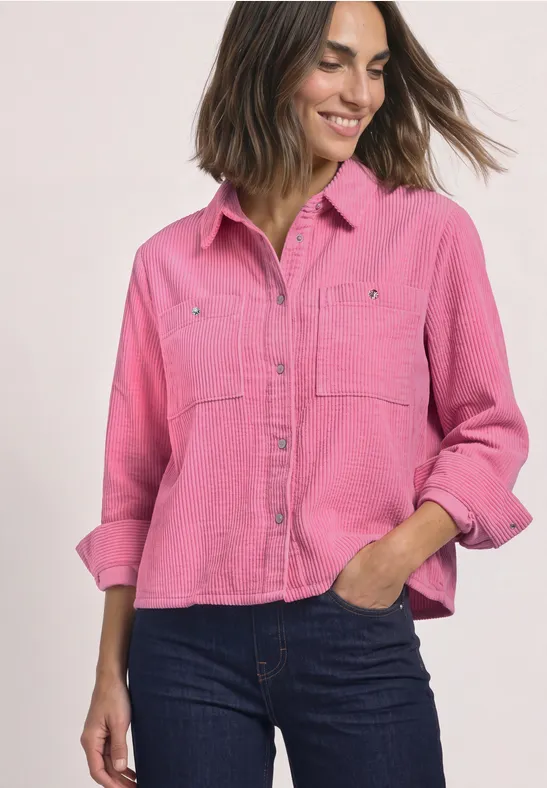Femme souriante en chemise rose décontractée aux manches retroussées et jean foncé.