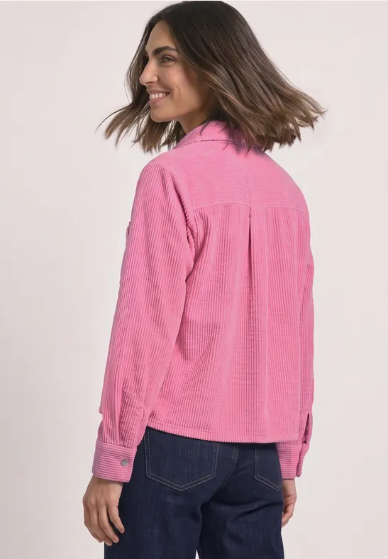 Une femme porte une chemise en velours côtelé rose et un jean foncé, vue de dos, souriante et regardant de côté.