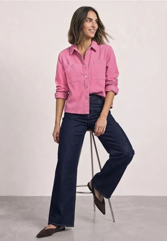 Femme en chemise rose et jeans larges bleu foncé appuyée de façon décontractée sur un tabouret.