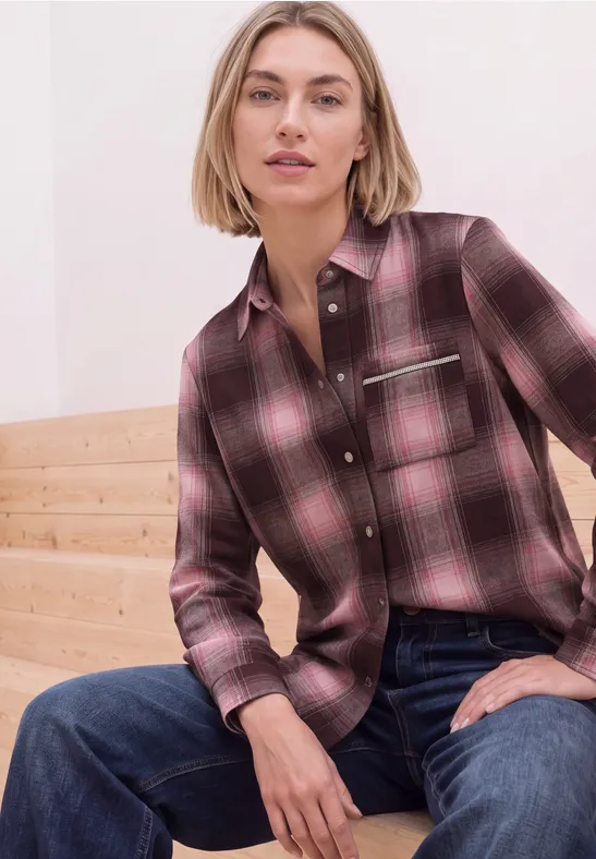 Vrouw in een roze geruite blouse en blauwe jeans zit ontspannen op lichte houten treden.
