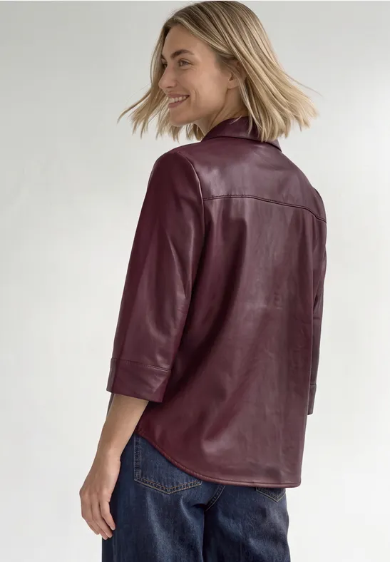 Vrouw draagt een bordeaux leren blouse en spijkerbroek, kijkt lachend over haar schouder.