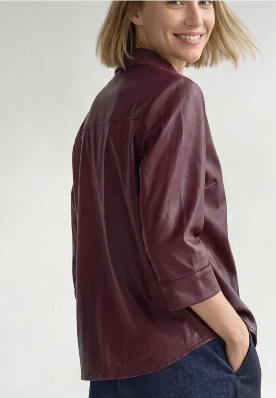 Vrouw in bordeaux leren blouse en spijkerbroek, handen in zakken, kijkt lachend over haar schouder op witte achtergrond.