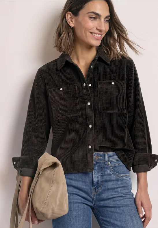 Vrouw glimlacht in zwart corduroy overhemd en blauwe jeans met een beige tas in de hand.