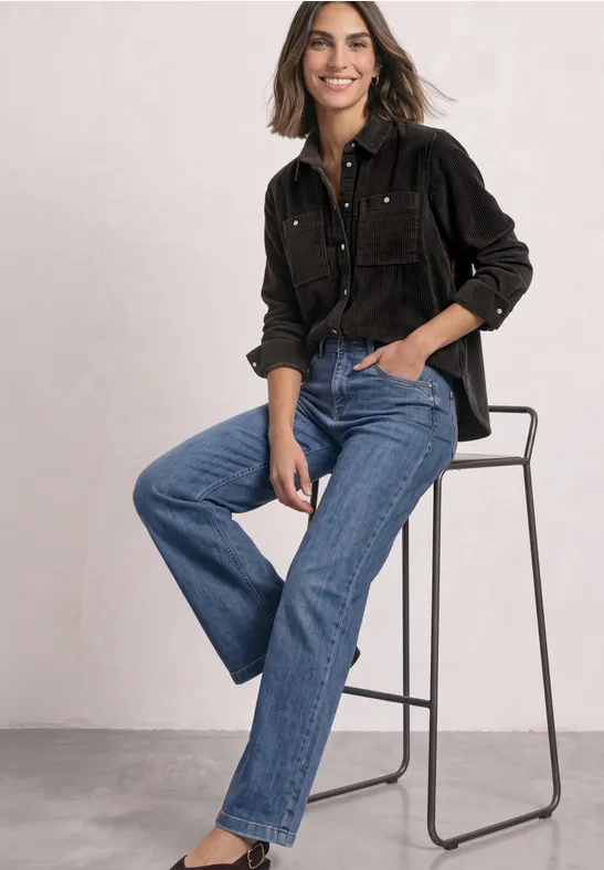 Vrouw zittend op hoge kruk in zwarte blouse en rechte blauwe jeans, ontspannen glimlachend.