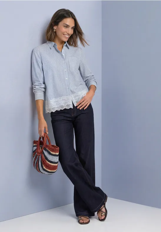 Femme en chemise bleu clair à dentelle, pantalon évasé sombre, sac rayé et sandales.