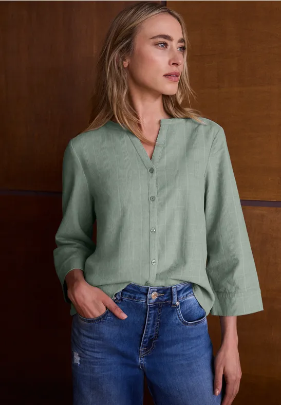 Vrouw met groene blouse en spijkerbroek leunt tegen een houten wand, kijkt naar rechts.