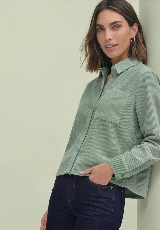 Femme portant un chemisier vert clair ample avec poche poitrine et un jean foncé.