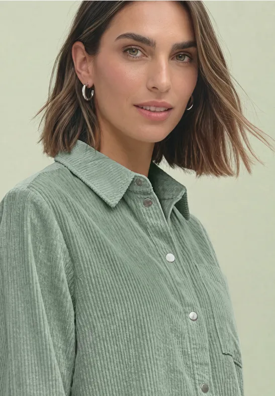 Portrait d’une jeune femme en chemise en velours côtelé verte et créoles argentées, léger sourire.