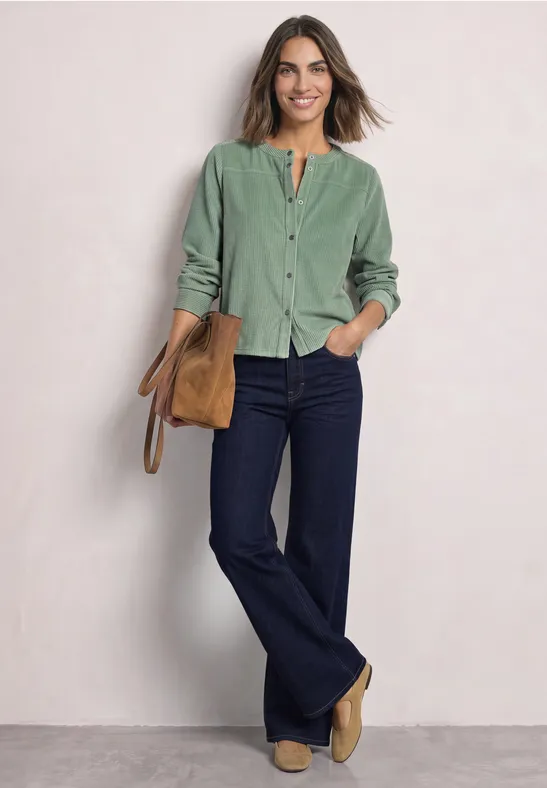 Femme portant une chemise en velours côtelé verte, jean flare foncé, bottines beiges et un sac devant un fond clair.