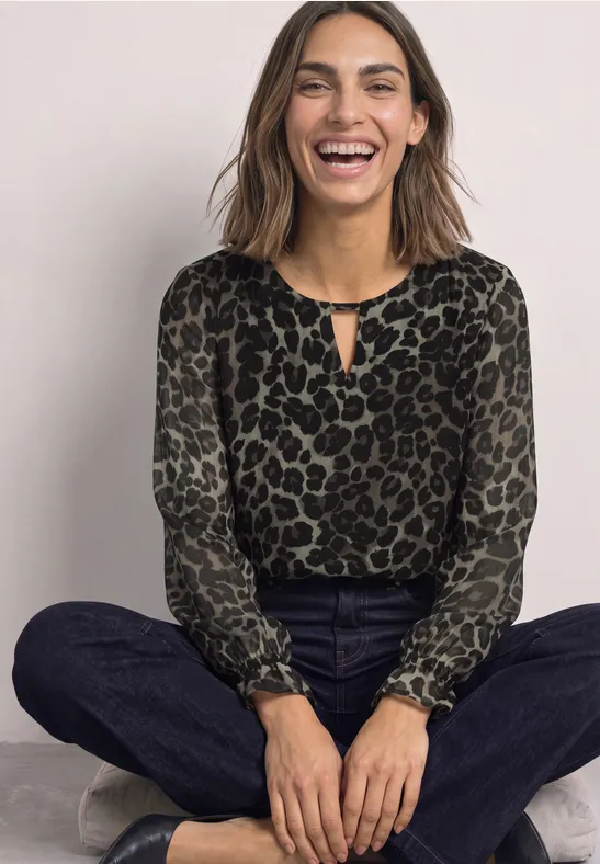 Lachende Frau sitzt crossbeinig in Leoparden-Print-Bluse, dunklen Jeans und Sneakers vor neutralem Hintergrund.